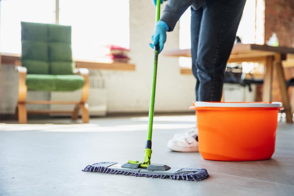 Person mopping a floor with a green mop and orange bucket in a bright, modern room.